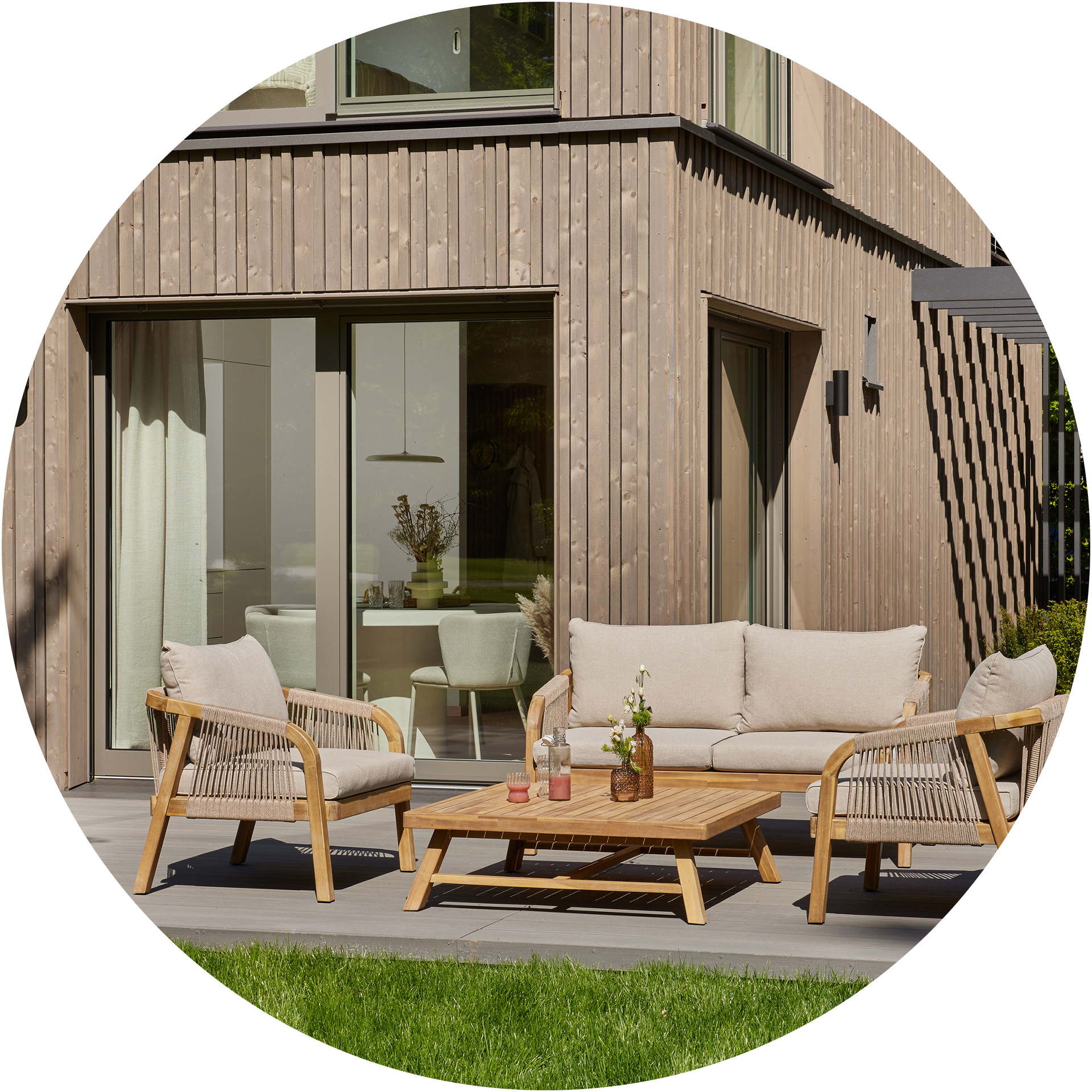 Ein moderner Außenbereich mit einem bequemen Sofa und zwei Stühlen aus natürlichem Holz. Auf einem Holzkaffee-tisch stehen Getränke und eine kleine Vase mit Blumen. Im Hintergrund sind große Fenster zu sehen, die einen klaren Ausblick auf das Innere des Hauses bieten.