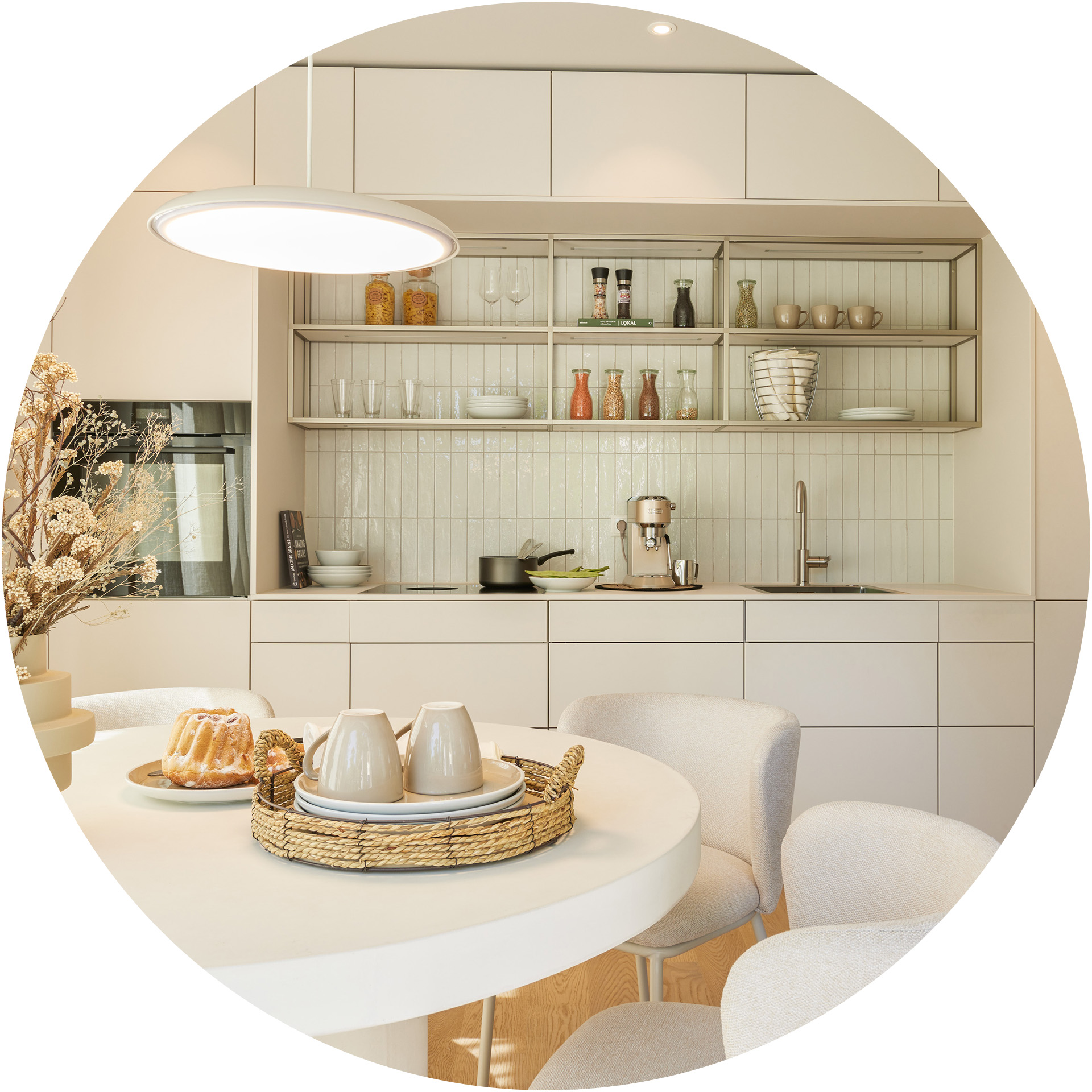 Eine moderne, helle Küche mit offenen Regalen, die verschiedene Gläser und Geschirr zeigt. Im Vordergrund ein runder Tisch mit einem Korb, der zwei Bechern und Tellern hält. Im Hintergrund steht eine Küchenmaschine neben einem Waschbecken.