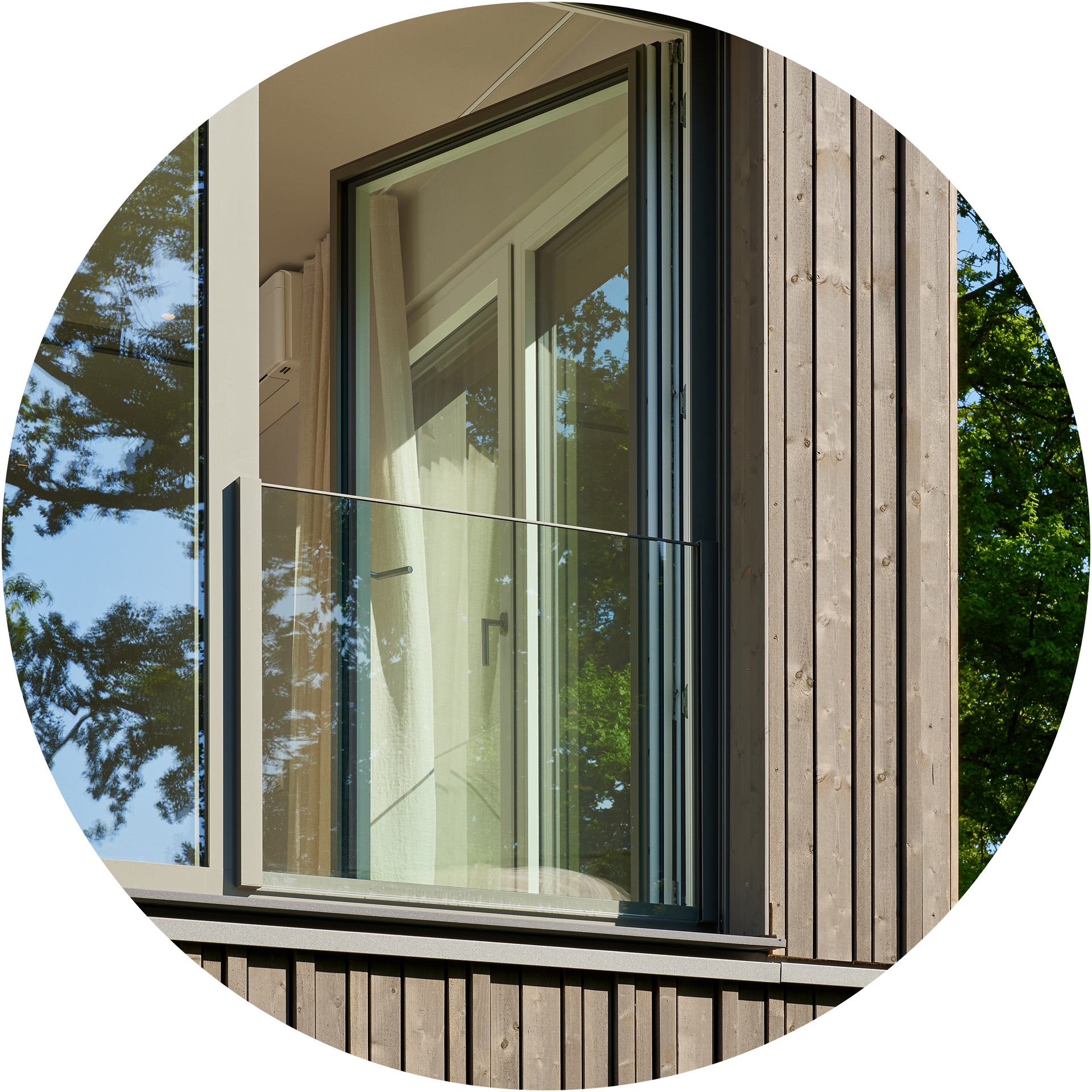 Ein modernes Fenster mit Glasschutzbalustrade in einem Holzgebäude. Durch das Fenster sind helle Vorhänge und ein Teil der Inneneinrichtung sichtbar. Im Hintergrund reflektieren sich Bäume auf der Fensteroberfläche.
