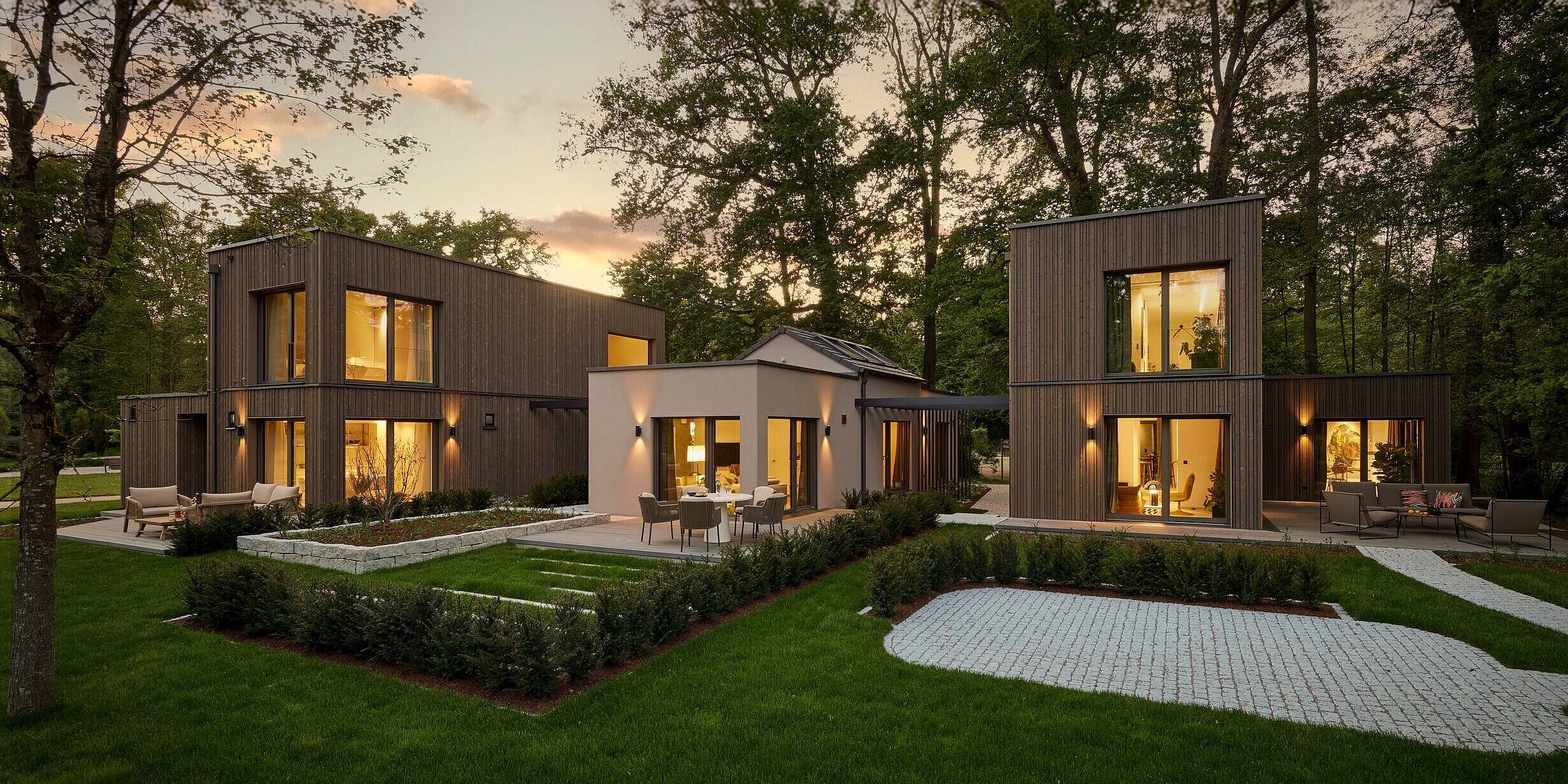 drei neue Ausstellungshäuser OPTION in der World of Living Moderne Häuser stehen in einer grünen Umgebung, umgeben von Bäumen. Die Gebäude haben große Fenster, durch die warmes Licht strahlt. Gepflasterte Wege und gepflegte Rasenflächen ergänzen die ansprechende Landschaft. Der Himmel zeigt sanfte Farben eines Sonnenuntergangs.