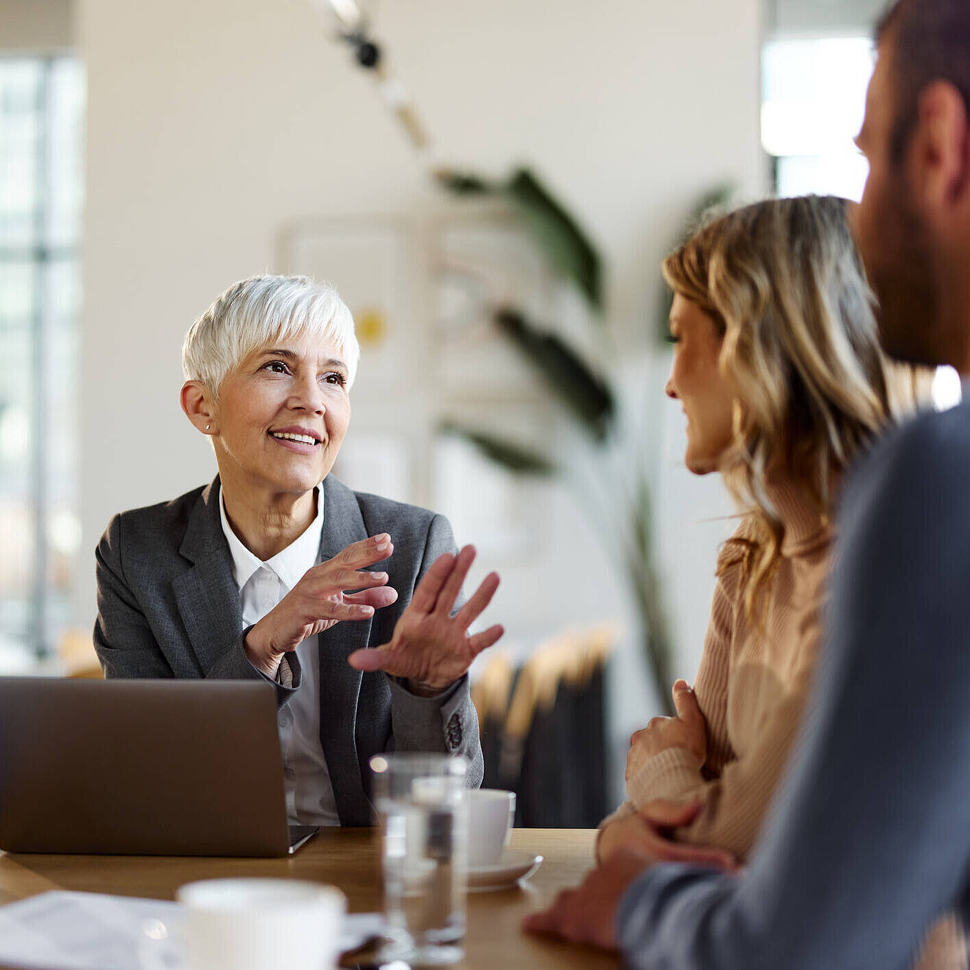 Beratungsgespräch in angenehmer Atmosphäre Eine Beraterin mit kurzem grauen Haar und einem freundlichen Lächeln spricht mit einem jungen Paar in einem modernen Büro. Vor ihr steht ein aufgeklappter Laptop, die Atmosphäre wirkt professionell und zugleich entspannt.